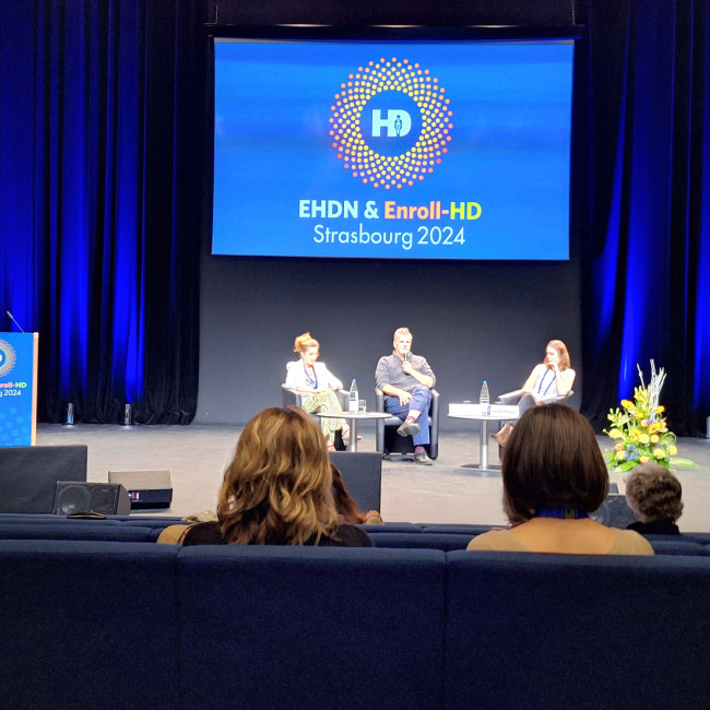 Three people sat on a stage, one holding a microphone in front of blue signage for the EHDN conference